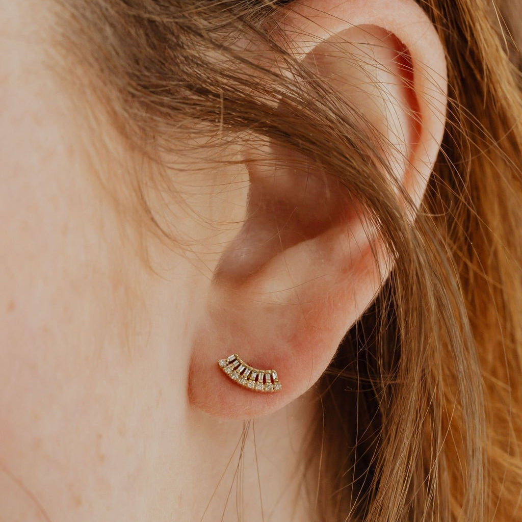 A close-up image of a woman's ear wearing a gold stud earring with a curved fan design set with diamonds.