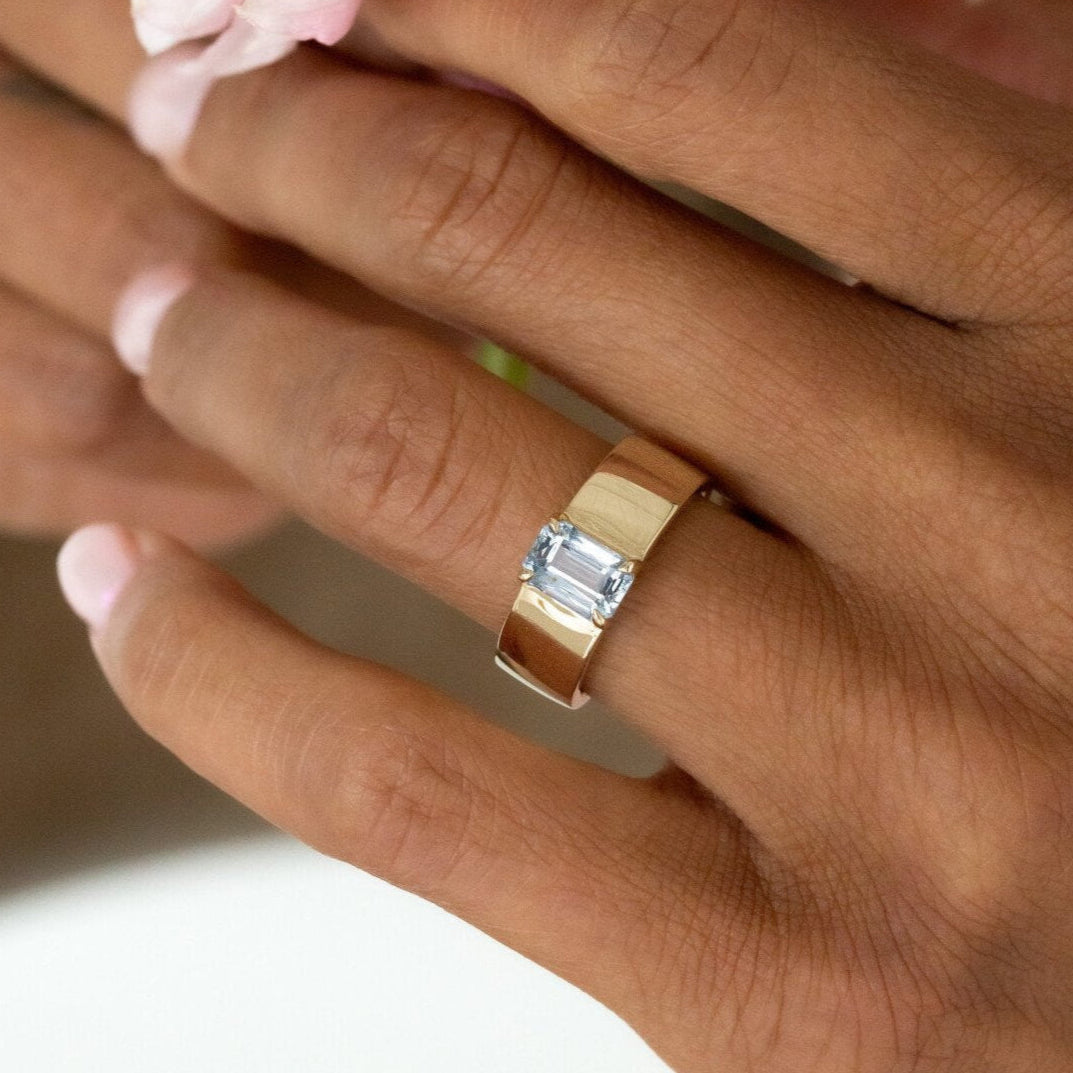 Hand wearing a gold ring with a blue sapphire, holding a pink flower.