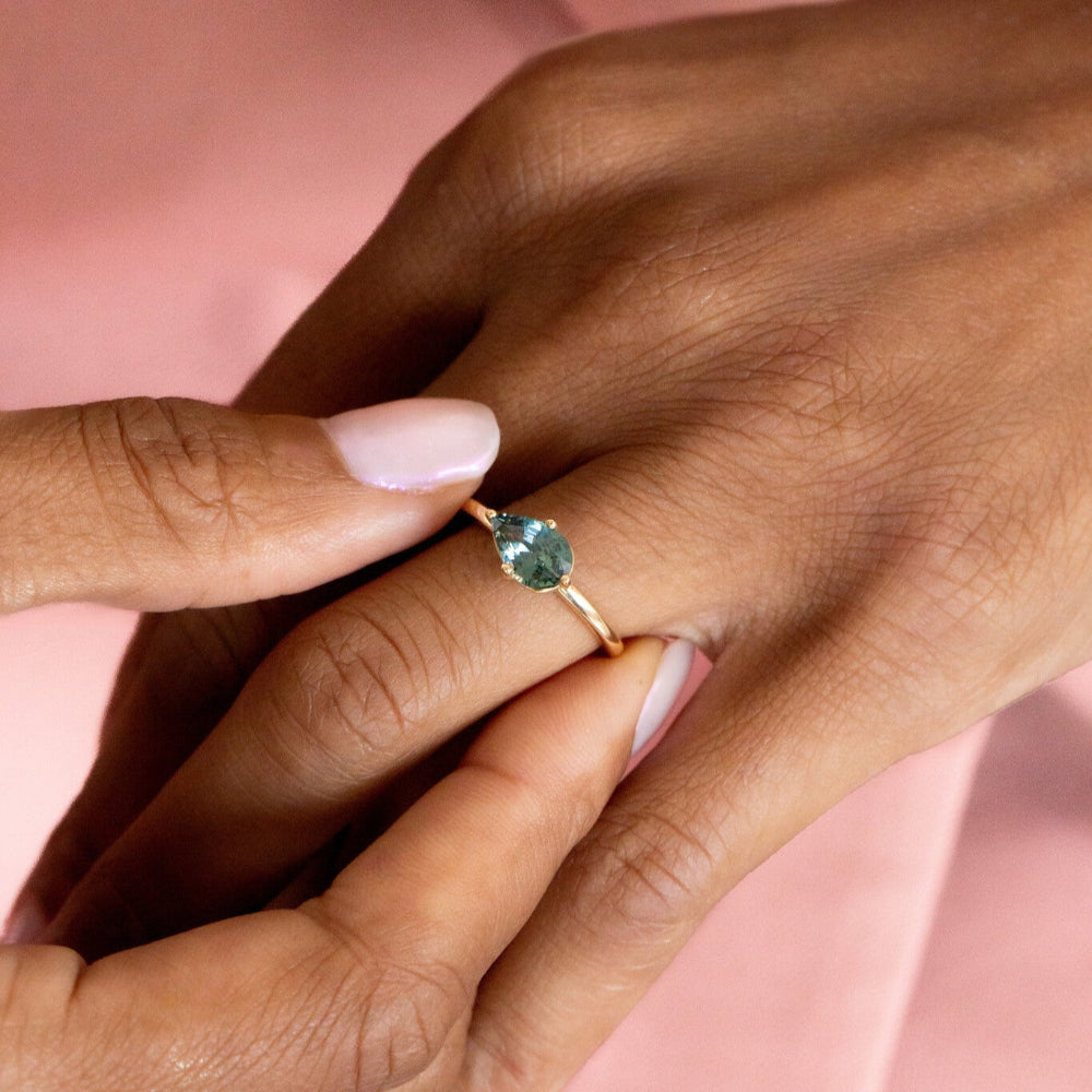 Close-up of a hand wearing a gold ring with a green sapphire gemstone on a pink background