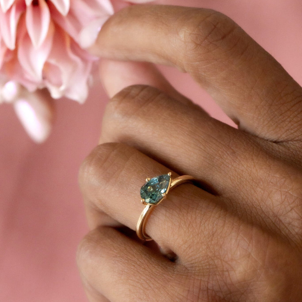 Hand wearing a gold ring with a green sapphire gemstone, holding pink flowers against a pink background