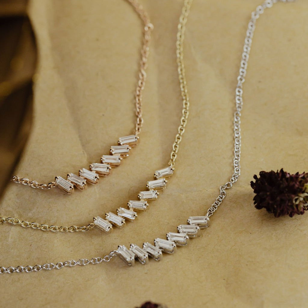 A curved bar necklace with baguette diamonds set in gold, displayed on a beige background with some floral elements.