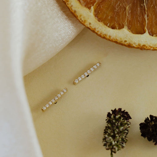 A pair of 14k yellow gold stud earrings with pave diamond bars, displayed on a white surface with a dried orange slice and a sprig of lavender.