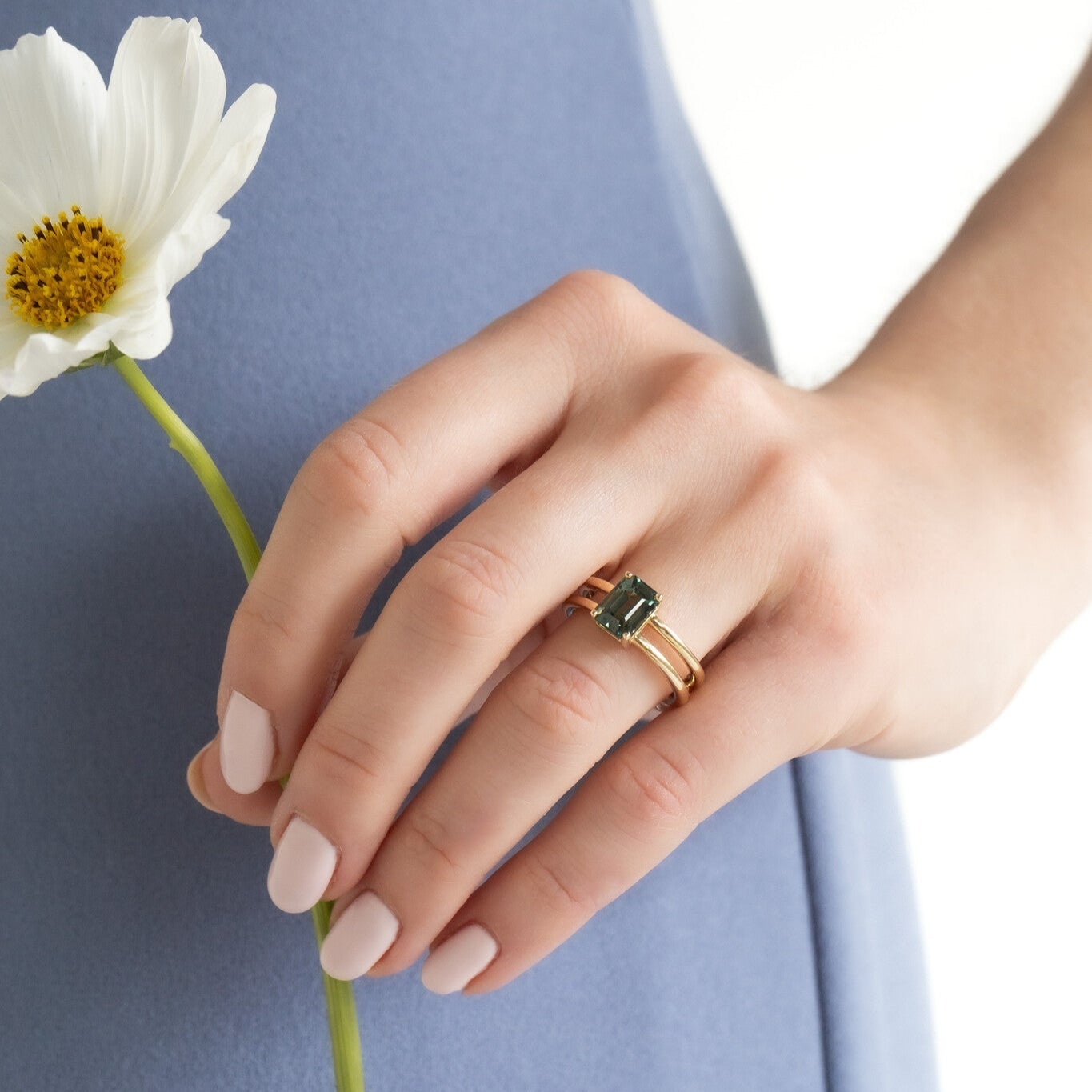 Hand wearing a gold ring with a green sapphire gemstone, holding a white flower against a blue background