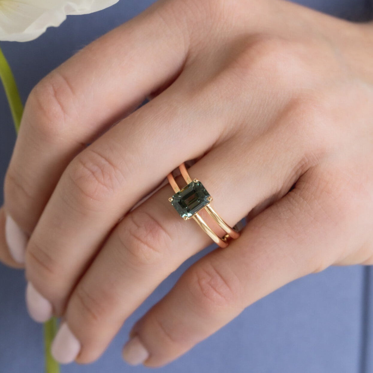 Hand wearing a rose gold ring with a green gemstone, holding a white flower against a blue background