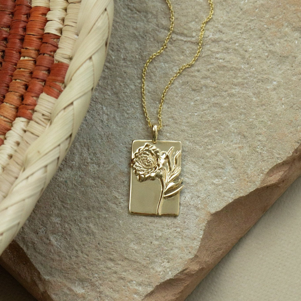 A gold necklace with a rectangular pendant featuring a dahlia flower design, displayed on a textured background.