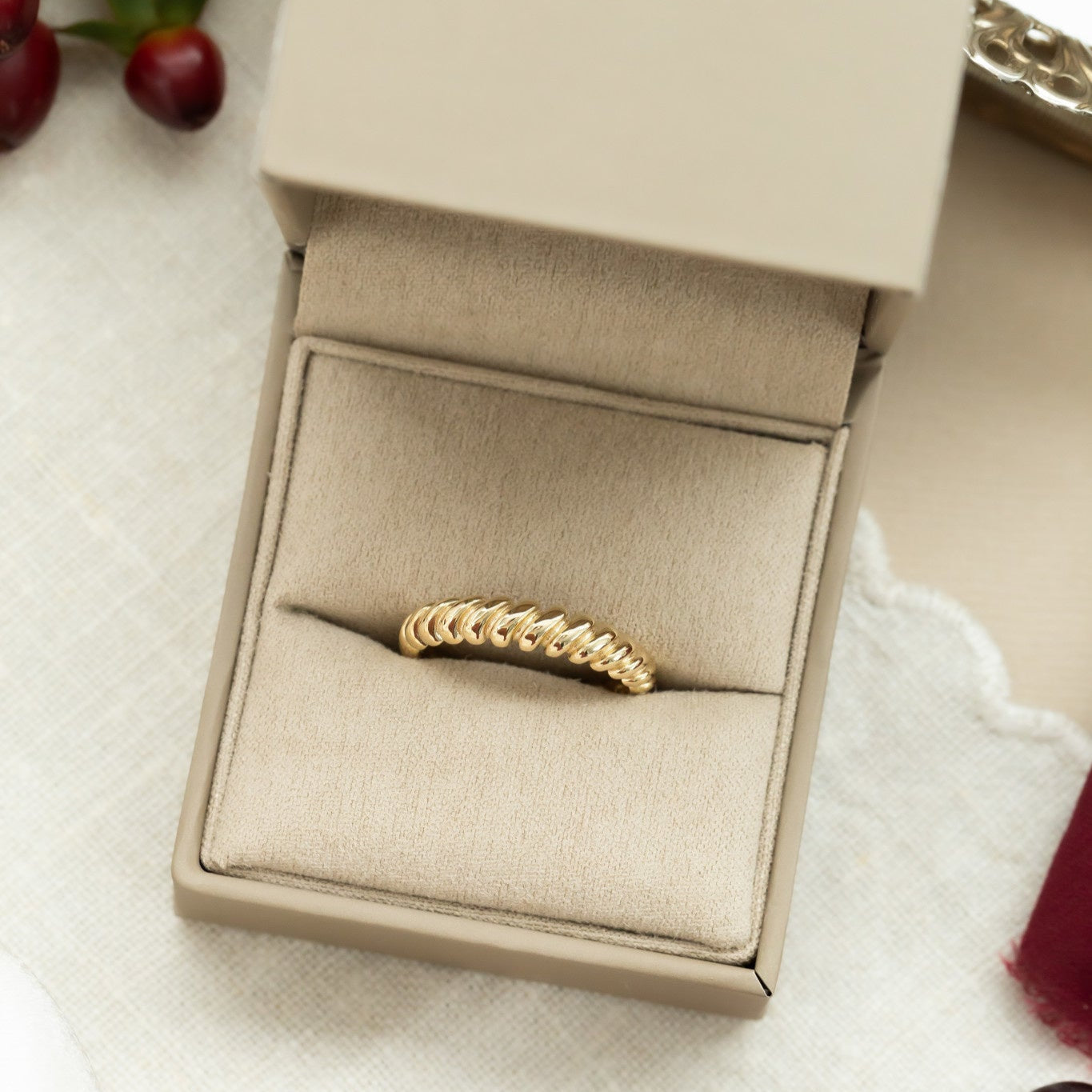 A 14k yellow gold ring with a rope texture on a 4.5 mm band, displayed in an open beige box, placed on a white fabric surface with some berries and a blurred background.
