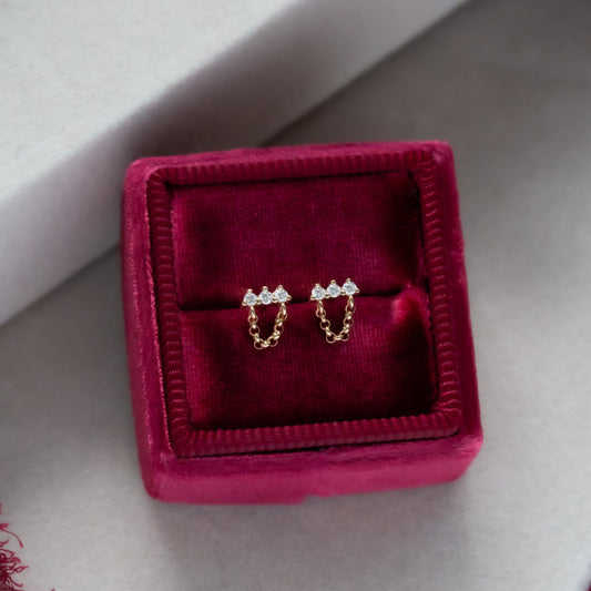 A pair of 14k yellow gold earrings with a diamond bar and three smaller stones, enclosed in a pink jewelry box with a soft interior.