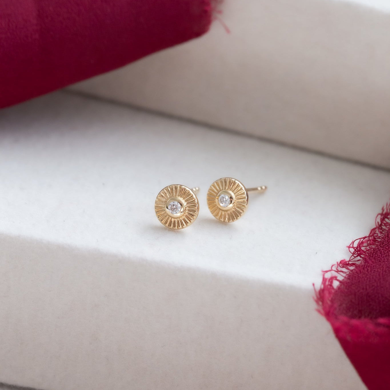 A pair of 14k yellow gold stud earrings with round textured disks and diamonds set in prongs, placed on a soft surface with a red cloth background.
