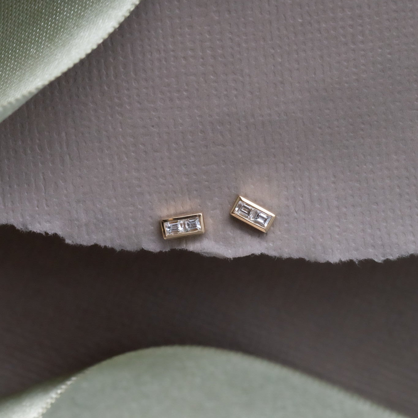 A pair of gold stud earrings with double baguette diamonds set in a high polish bezel, displayed on a green fabric surface.