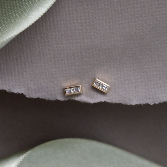 A pair of gold stud earrings with double baguette diamonds set in a high polish bezel, displayed on a green fabric surface.