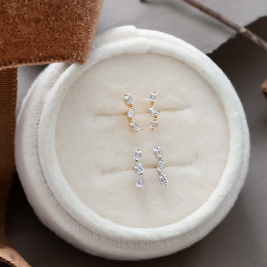 14k gold stud earrings with .21 ct baguette diamonds in a zig zag design, set in prongs, displayed on a soft surface with brown leaves around.