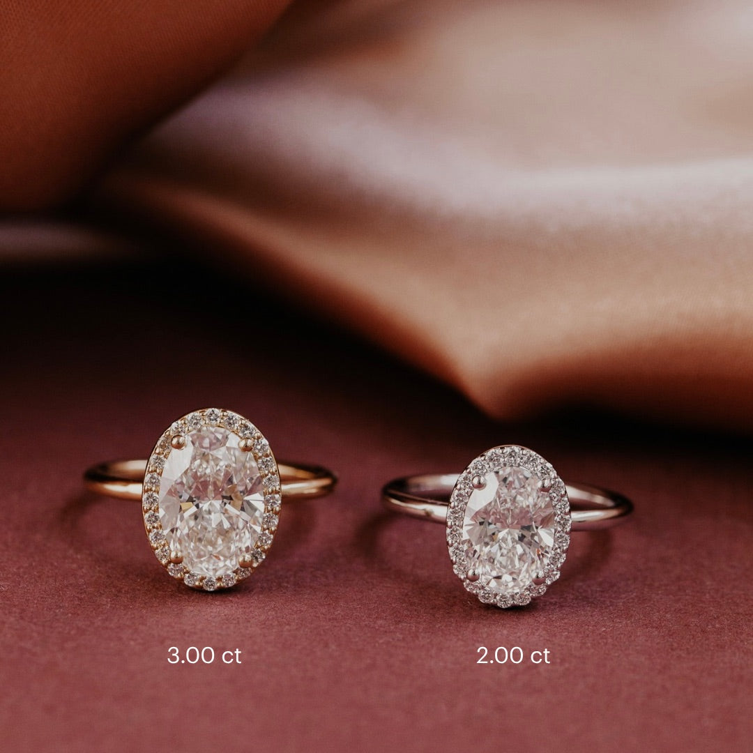 The Audrey Oval Ring in 14k yellow gold and white gold shown in two center stone size options: 3.00 ct (left) and 2.00 ct (right), displayed on draped pink satin fabric.

