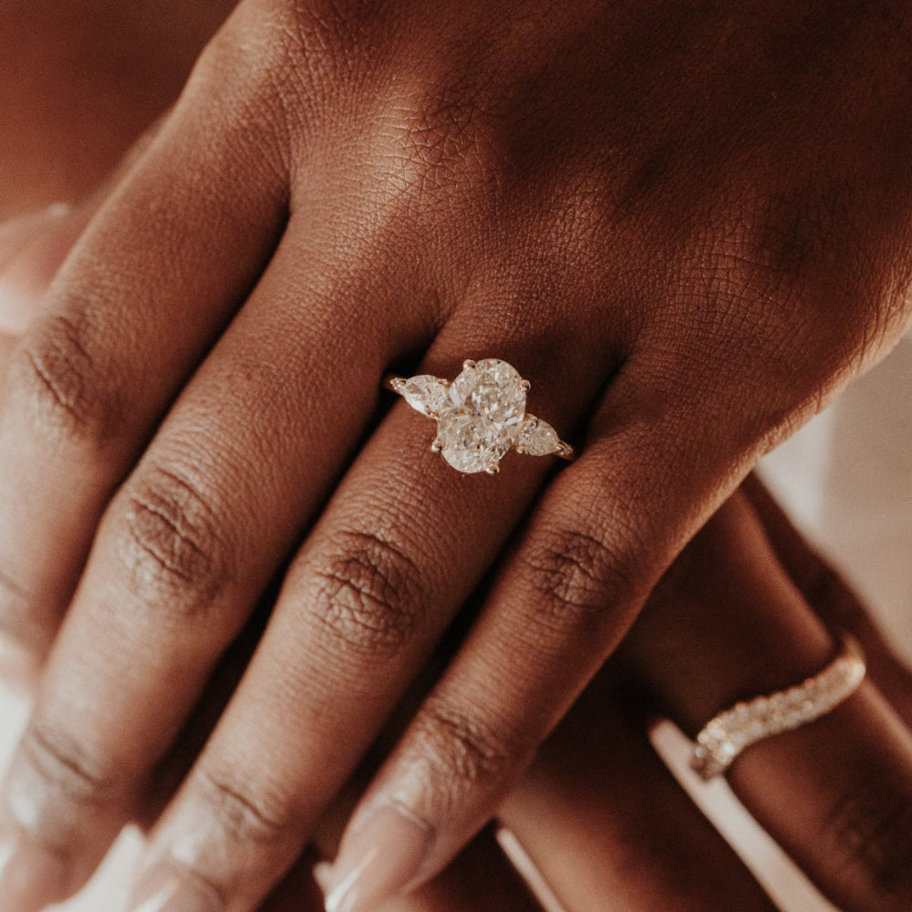 Close-up of a hand wearing the Toula oval diamond engagement ring with diamond side stone on a blurred backgroun