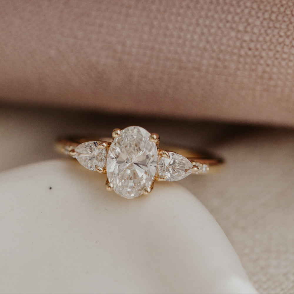 Close up of the Toula diamond engagement ring on a white surface with a pink fabric background.