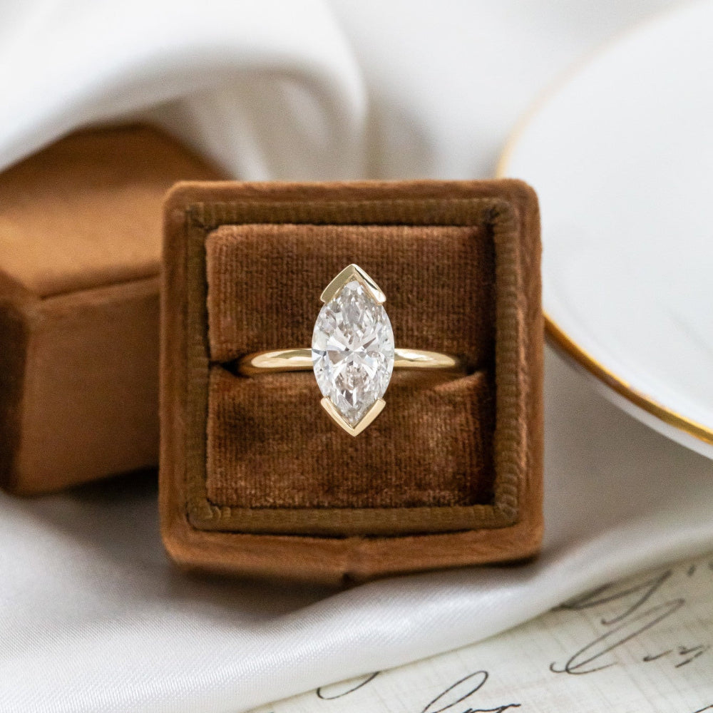 Gold Zadie engagement ring with a marquise diamond in a brown velvet box on a white surface.