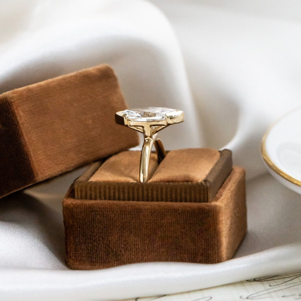 Side view of the gold Zadie engagement ring with a marquise diamond in a brown velvet box on a white fabric background.