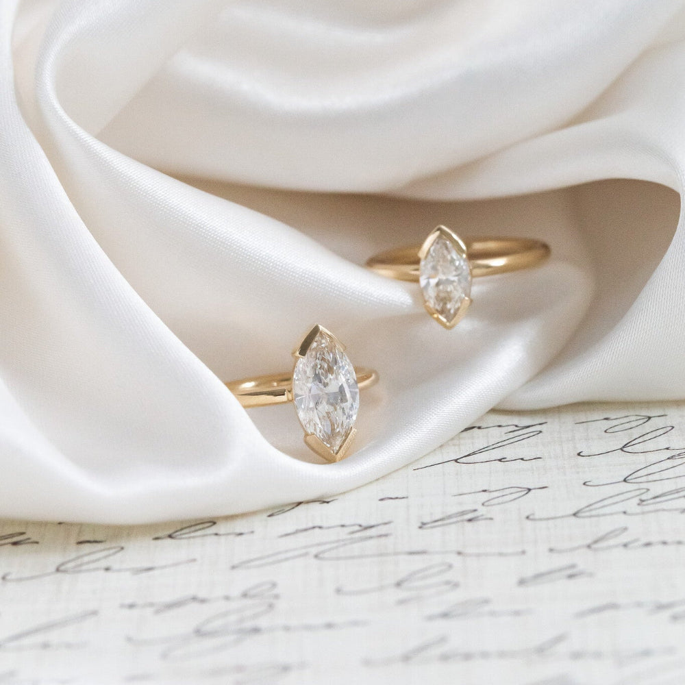 Two gold Zadie engagement rings with marquise-shaped diamonds on a white fabric background.