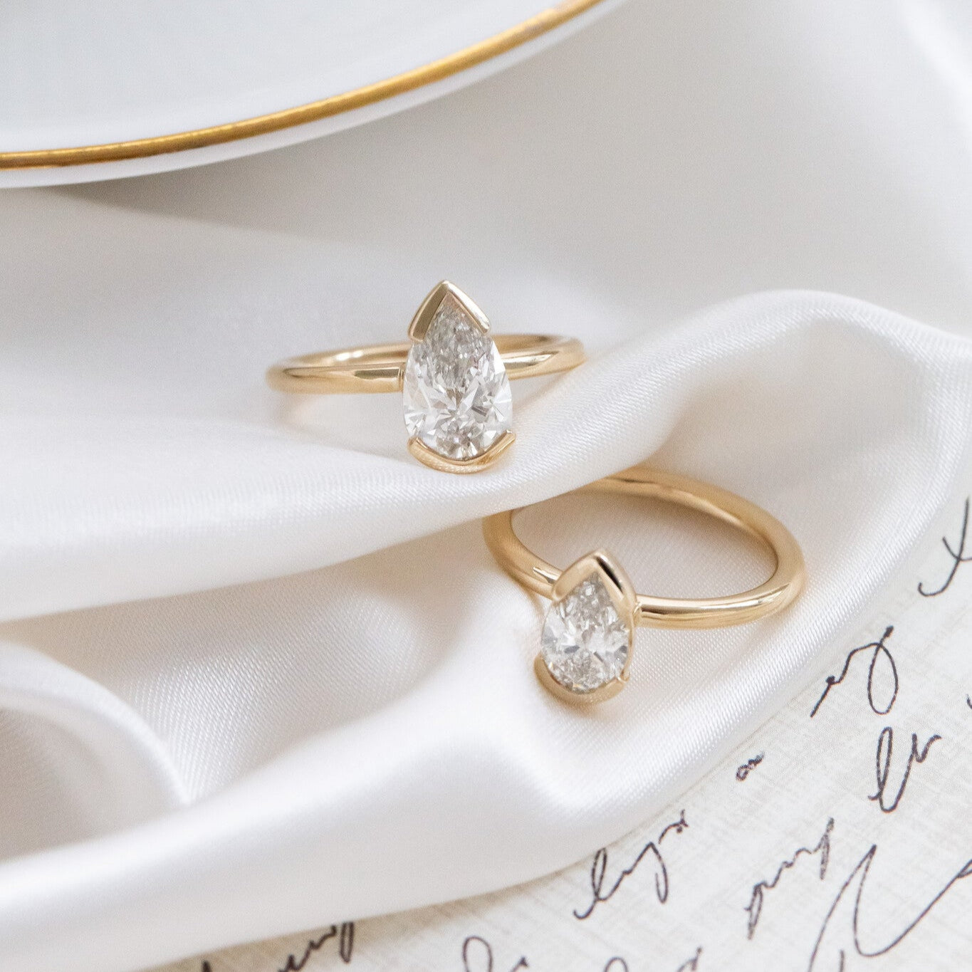 Two Zadie pear-shaped solitaire rings with a white diamonds, set in a half bezel design, placed on a white surface with a gold-rimmed dish in the background.