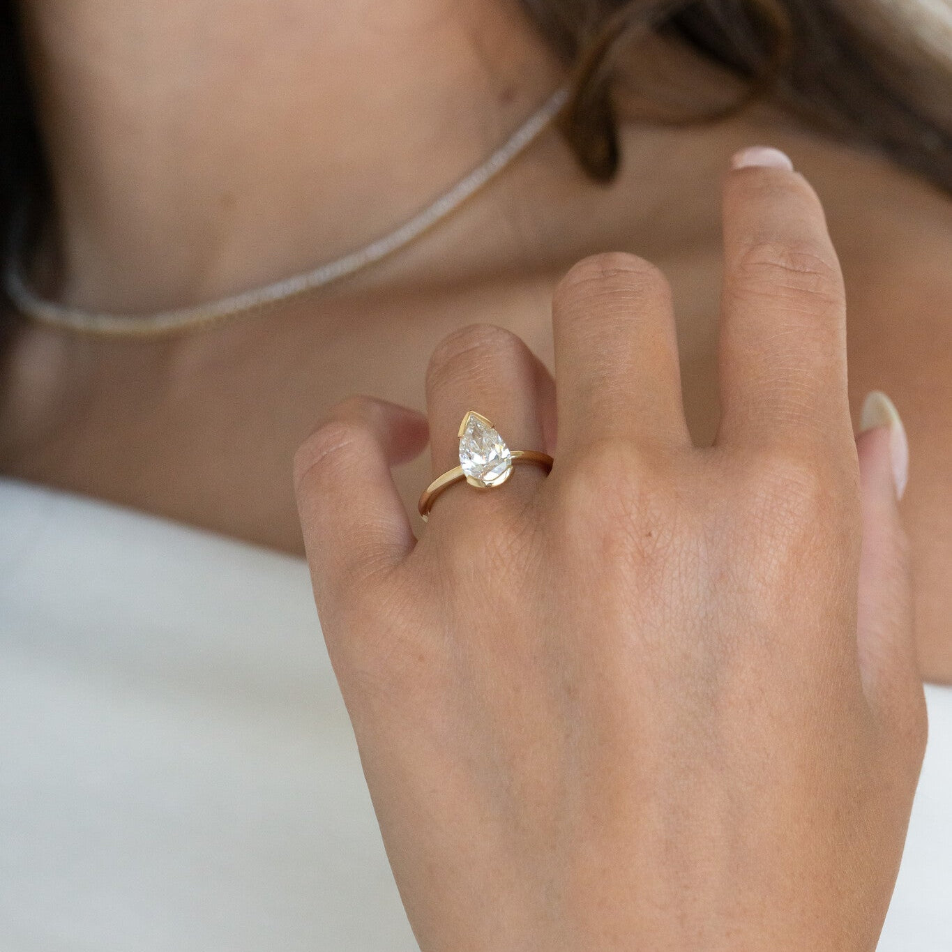 Close-up of a hand wearing a gold Zadie solitaire engagement ring with a pear-shaped diamond on a neutral background.