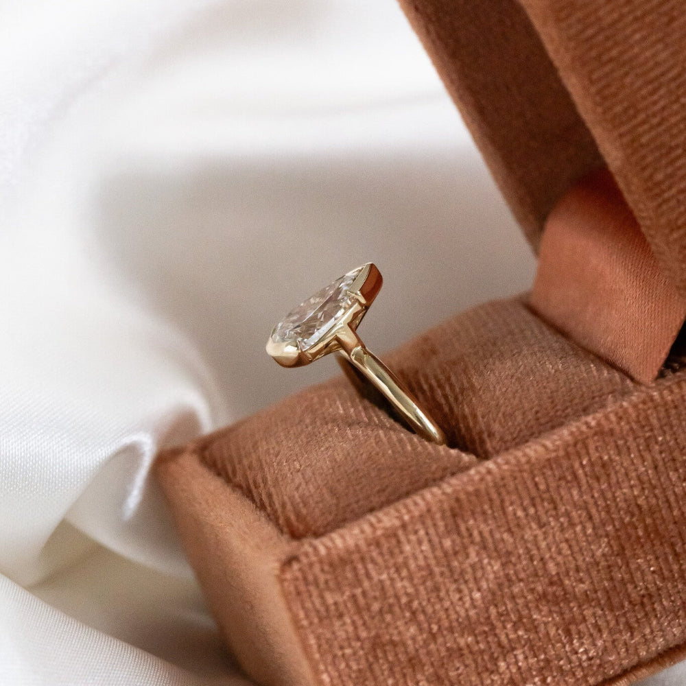 Gold Zadie engagement ring with a pear-shaped diamond on a brown fabric background.