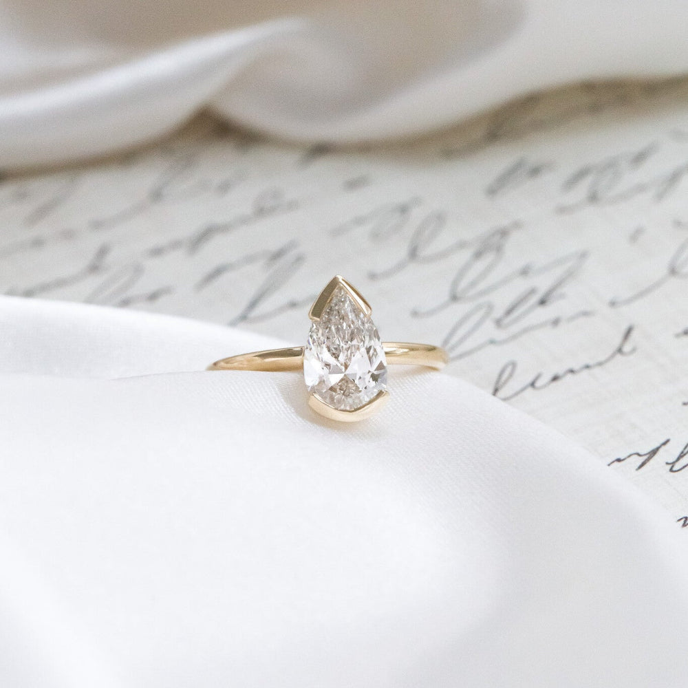 Gold Zadie engagement ring with a pear-shaped diamond on a white fabric background.