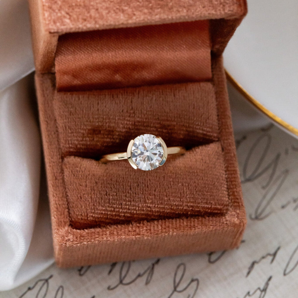 A Zadie round solitaire ring with a white diamond, set in a half bezel design, placed in a brown velvet box.